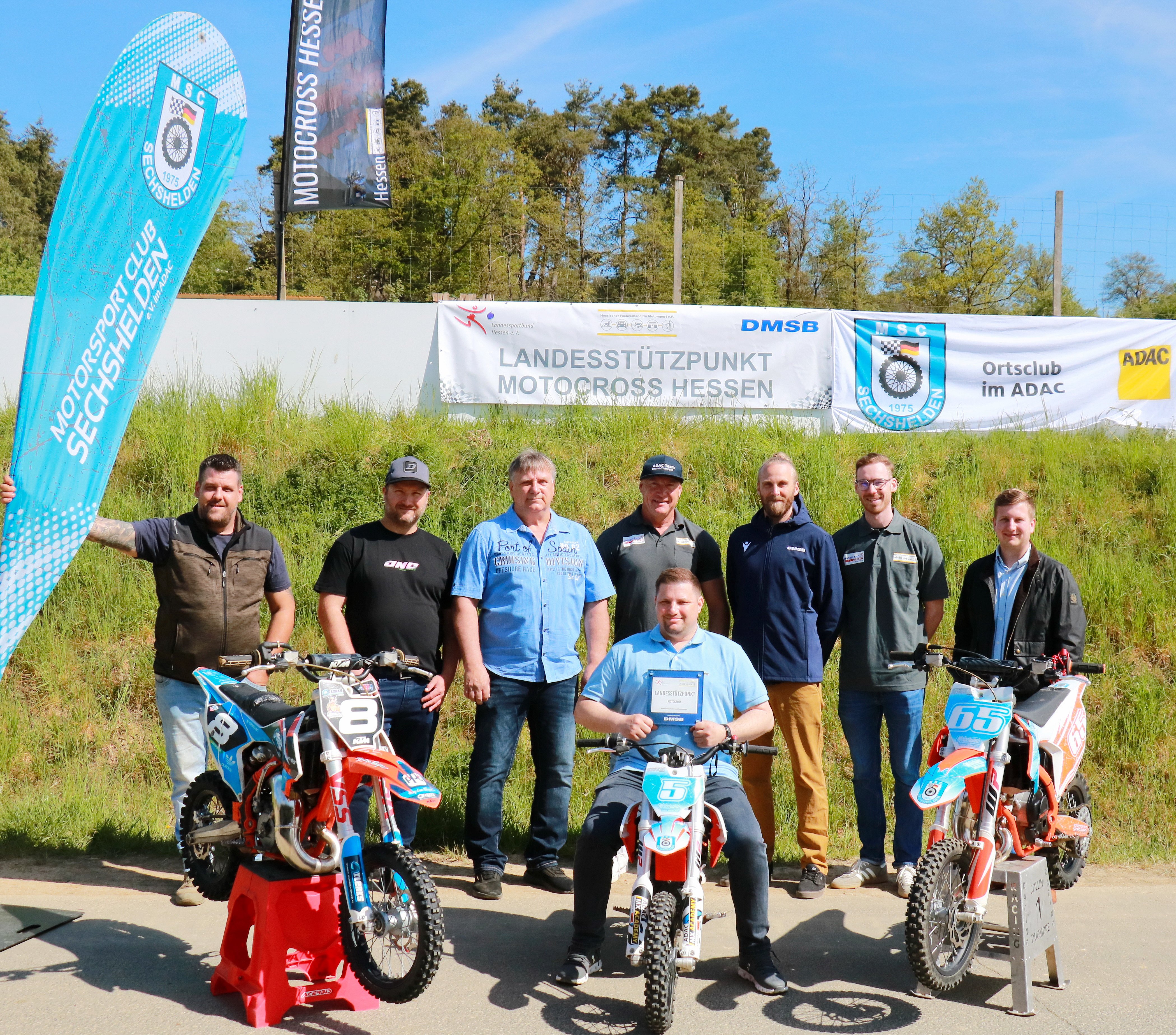 Acht Männer stehen an einem sonnigen Tag hinter drei Motocross-Motorrädern mit Bannern und Fahnen im Hintergrund.